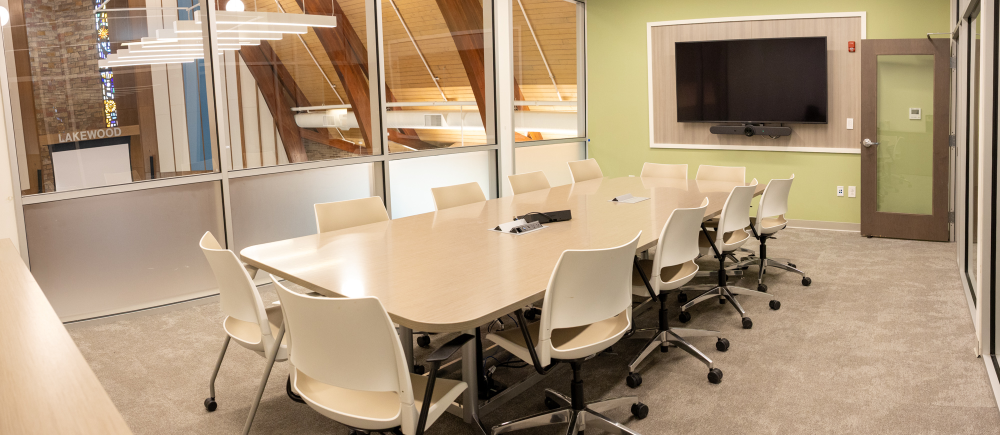 Second floor conference room at Cove Community Center. A large conference table with 12 chairs, windows on the back wall, a TV screen mounted on the right wall. Carpeted floor.